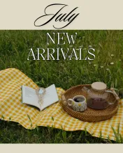 July New Arrivals. Photo of an open book and coffee on a yellow gingham picnic blanket outdoors.